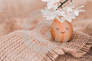 Creative easter egg in floral wreath, cute face and sleepy eyes on sackcloth background. Happy Easter concept