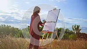 Creative female woman with a brush and palette in her hands draws a picture on canvas using an easel in nature against