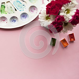 Creative flat lay of watercolor palette, watercolor cuvettes and beautiful bouquet of flowers. Artist workplace on a pink pastel b