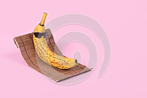 Creative fun idea with a banana in sunglasses lying on a sun bed on a pastel pink background