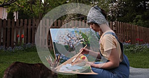 Creative girl is painting a beautiful picture in the garden, holding a palette and brush, using tools to create a canvas