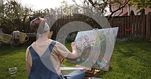Creative girl is painting a beautiful picture in the garden, holding a palette and brush, using tools to create a canvas