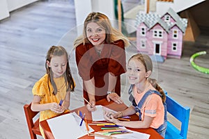 Creative kids during an art class in a daycare center or elementary school classroom drawing with female teacher.