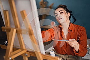 A creative Latin artist is working on a new painting. Happy young lady smiles cheerfully while painting on canvas in her