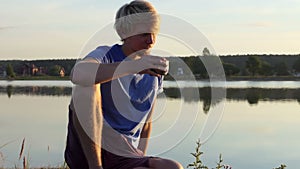 Creative man drinks coffee sitting on a lake bank at sunset in slo-mo