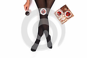 Creative photo of female legs in black pantyhose with a cup of coffee and a box of small cakes on a white background. Concept.