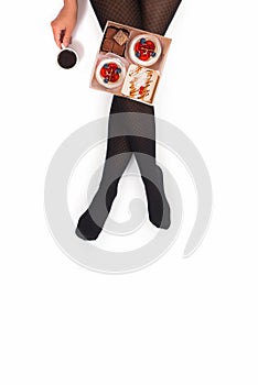 Creative photo of female legs in black pantyhose with a cup of coffee and a box of small cakes on a white background. Concept.