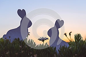 Creative photo of two silhouette paper rabbits in the chamomile flowers and green grass on the sunset sky background