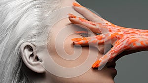 Creative portrait of a beautiful girl with hand on face, white hair. The hand is painted with orange paint.