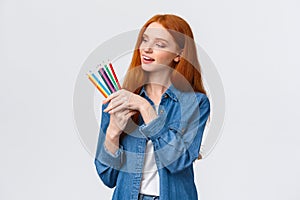 Creative and skilful good-looking redhead female in denim shirt, picking colored pencils, smiling thinking what draw