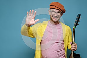 Creative smiling man in bright clothes with acoustic guitar makes a welcoming hand gesture and looks at the camera. Copy