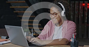 Creative young African American woman listening to music with headphones dancing and working with laptop in office