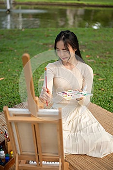 A creative young Asian woman is painting or drawing on a canvas while relaxing in a green park