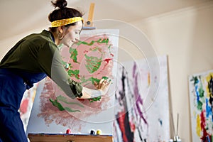 Creative young woman in apron painting a picture on easel, applying paint on canvas with fingers, standing in living