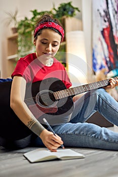 Creative young woman in casual wear making notes in her notebook while playing acoustic guitar, studying music at home