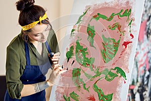 Creative young woman occupied in painting abstract picture, applying paint on canvas with fingers, standing in living