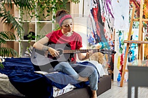 Creative young woman playing guitar while sitting on a bed in modern studio apartment