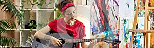 Creative young woman playing guitar while sitting on a bed in modern studio apartment