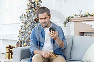 Credit score problems. A mature man is sitting at home on the sofa on Christmas holidays and holding a credit card in