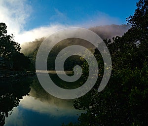 Beautiful view of creek with reflections blue sky, light clouds, fog, mountains and trees on water