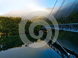 Beautiful view of creek with reflections suspension bridge, blue sky, light clouds, fog, mountains and trees on water