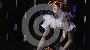 Creepy jester girl in a dress and a red hat with little bells is making faces and shaking her head