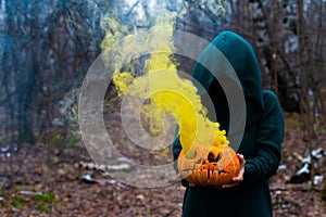 A creepy witch holds a steaming pumpkin in a deep forest. Jack o lantern emits yellow smoke for halloween