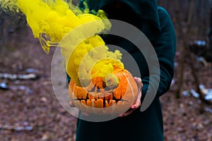 A creepy witch holds a steaming pumpkin in a deep forest. Jack o lantern emits yellow smoke for halloween