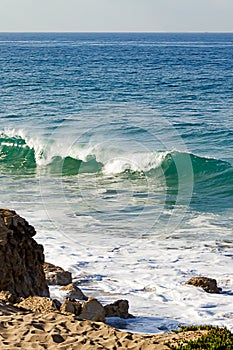 Cresting breaking wave on shoreline edge with washback foam on sandy shoreline with boulders