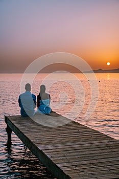 Crete Greece, young romantic couple in love is sitting and hugging on wooden pier at the beach in sunrise time with