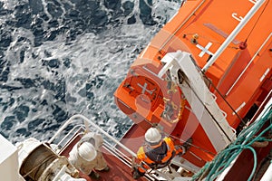 Crew member with orange jacket and helmet checking aft hook on life boat fixed to container vessel, second seaman in white overall