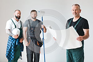 Crew of three builder with painting roll and renovation plans standing in empty interior