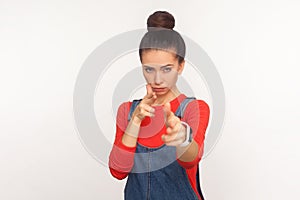 Crime, killing gesture. Portrait of dangerous girl with hair bun in denim overalls imitating guns, shooting into camera