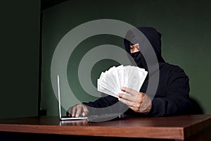 Criminal man in black hood holding money banknotes mockup on hand, looking at laptop computer at desk, mask thief robber