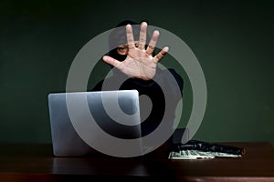 Criminal man in black hood with laptop computer, a gun and money at the at desk reaching out his hand to camera , mask thief