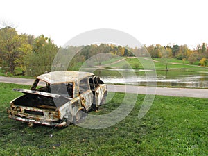 Criminals burned the car and left at a river. Burnt car on the grass by the pond. Damaged and abandoned vehicle