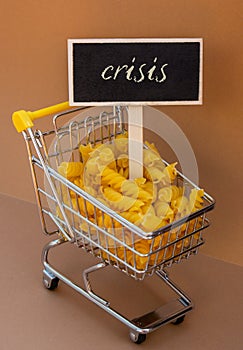 CRISIS text on Blackboard label against Shopping trolley cart Filled With Pasta on Beige background. Food and groceries