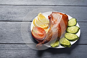 Crispy chicken legs, roasted on a grill with sliced lemon, cucumber and tomatoes on white plate on a wooden table. Copy space