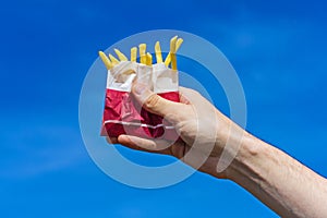 Crispy french fries in a paper bag in a male hand on a blue sky background.