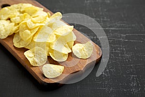 Crispy potato chips with salt on a rustic wooden board on a dark surface, side view. Space for text