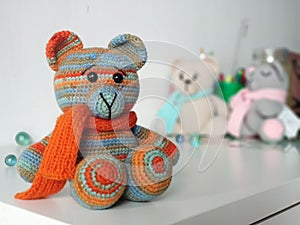 Crochet bear is orange-blue in scarf on background of other toys on light background. Knitted soft toy Teddy