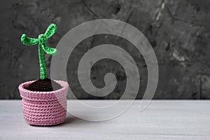 Crocheted green decorative flower with thee leaves in pink cocheted pot stands on light table.Blak grey background of concrete.