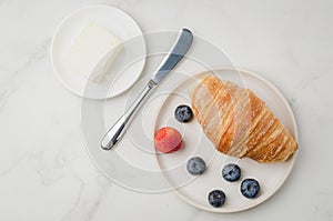 Croissant with berries in white bowl and butter knife on white stone background. Top view