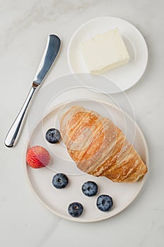 Croissant with berries in white bowl and butter knife on white table. Top view. Healthy breakfast