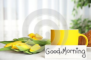 Croissant, coffee, flowers and card with words GOOD MORNING on white wooden table indoors