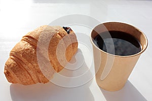 Croissant and coffee to go in brown craft paper cup on white background.