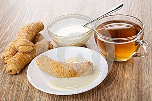 Croissant with condensed milk on saucer, croissants, spoon in bowl with milk, cup of tea on table