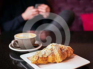 Croissant with flaked almonds on a white plate and a cup of coffee on the table in the cafe and a person holding smartphone on the
