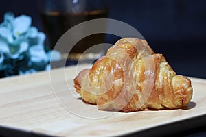 The croissant placed on the square wooden tray with out focus tea in a glass and blue flowers on dark background