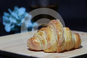 The croissant placed on the square wooden tray with out focus tea in a glass and blue flowers on dark background